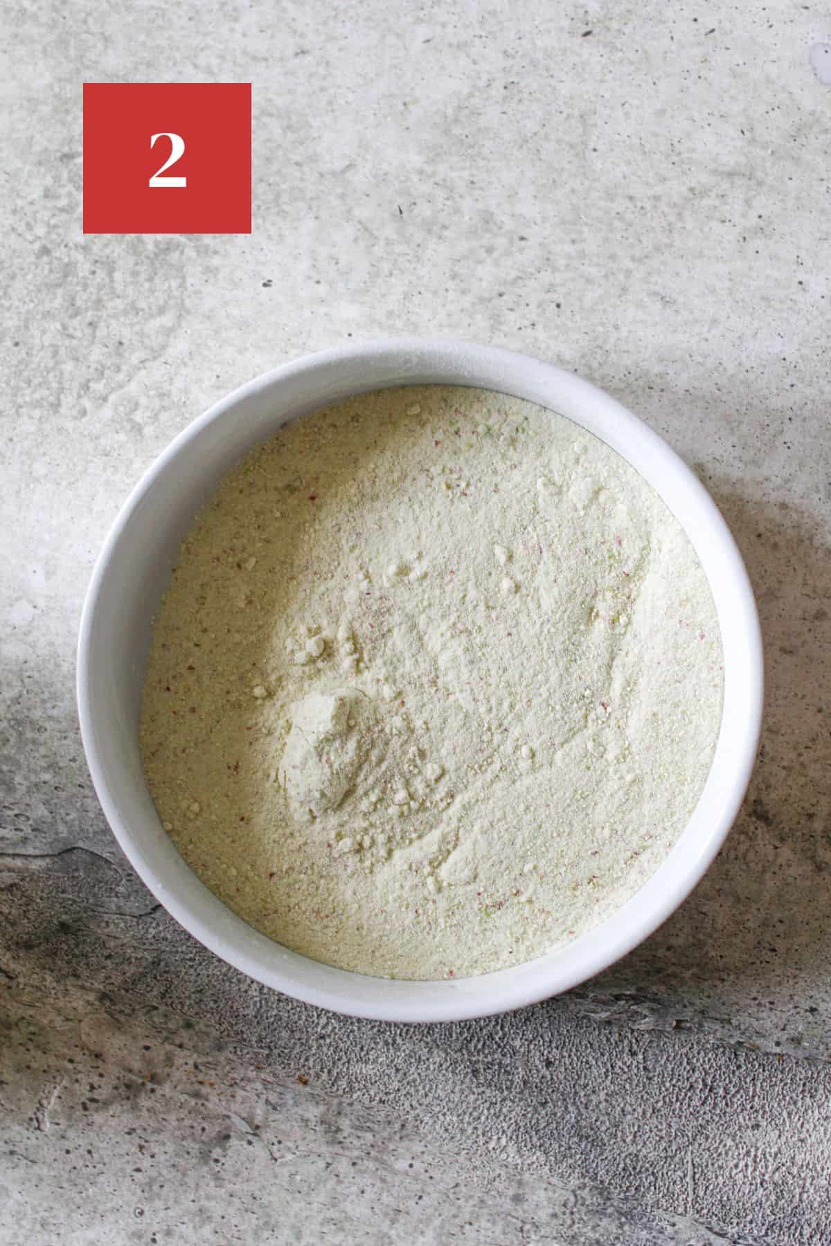 A white bowl with freeze dried apple powder on a cement background. In the upper left corner is a dark red square with a white '2' in the center.