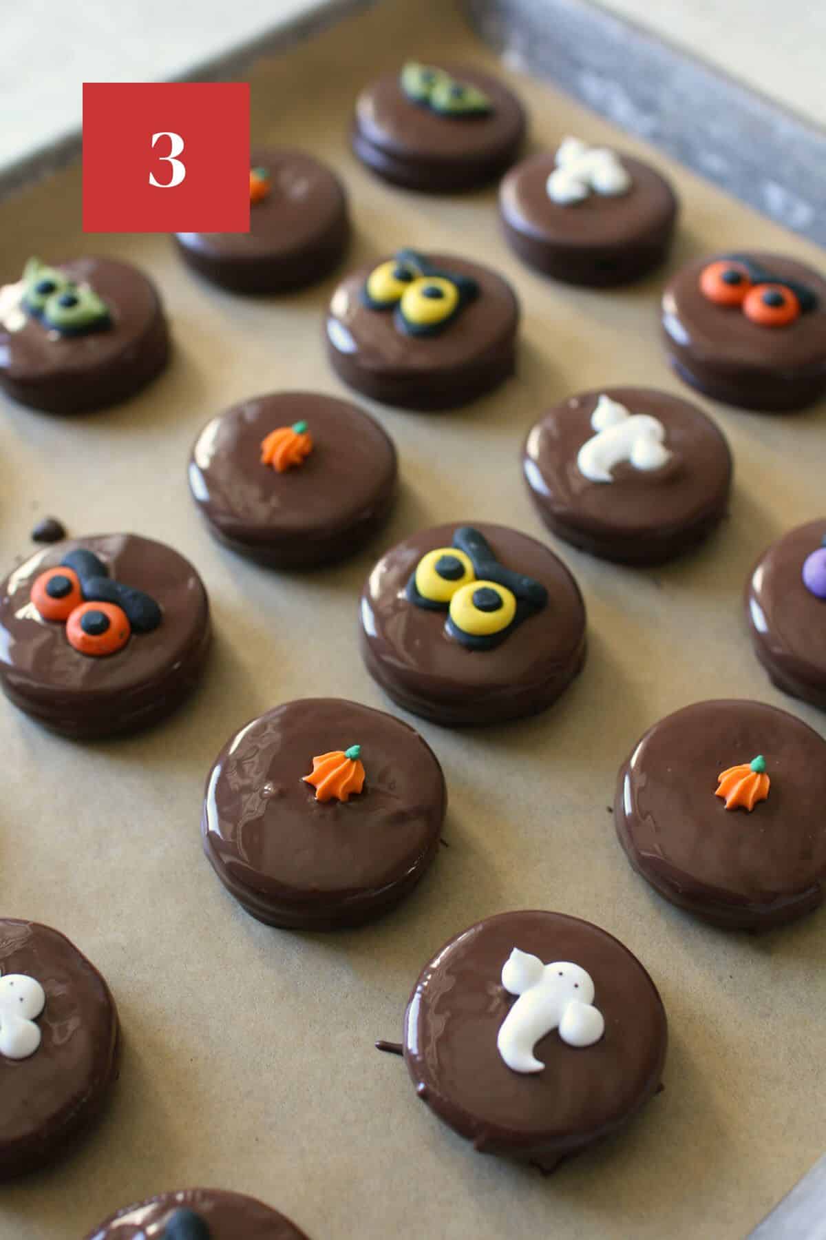 Dark chocolate covered oreos decorated with a halloween royal icing decoration on a piece of brown parchment paper on a baking tray on a cement background. In the upper left corner is a dark red square with a white '3' in the center.