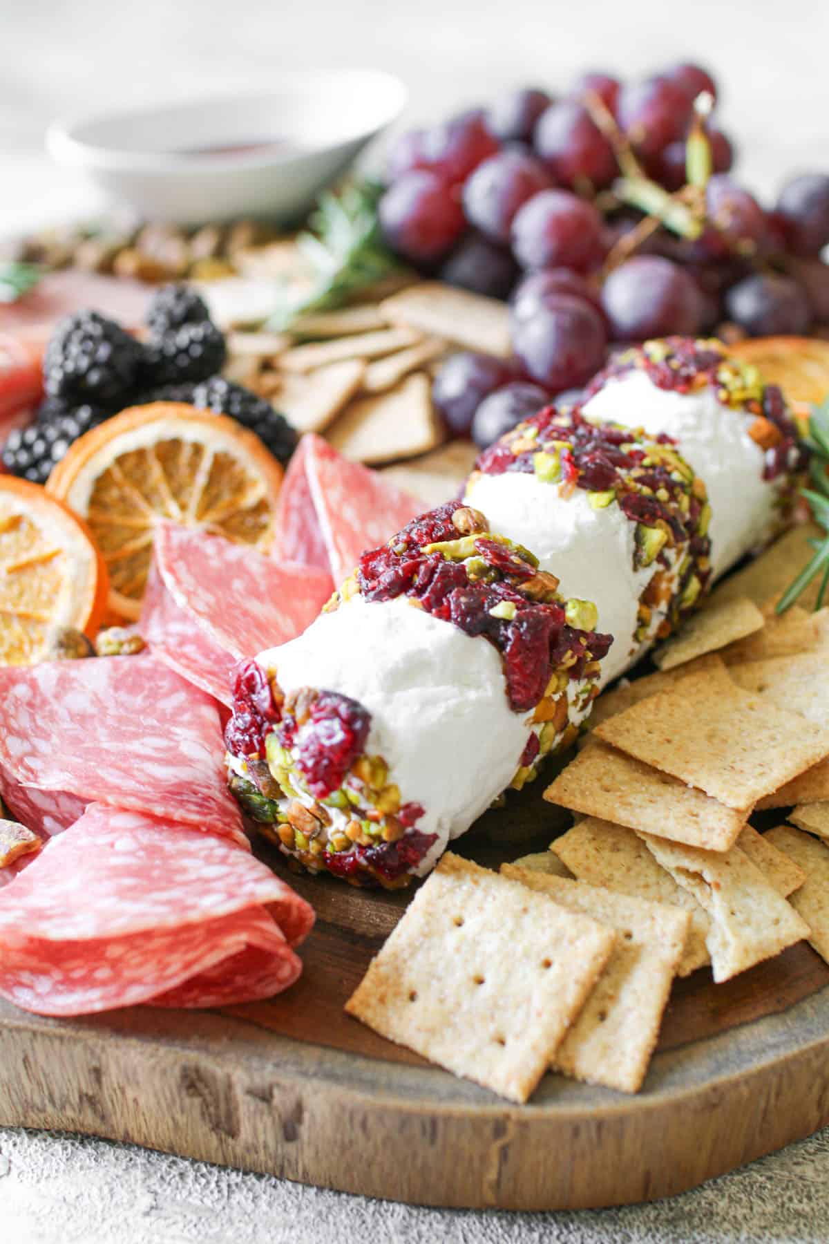 Close up of a Cranberry Pistachio Cheese Log surrounded by crackers, grapes, meats and garnished with dried oranges and fresh rosemary. The charcuterie sits on a wood truck board and sits on a stone background.