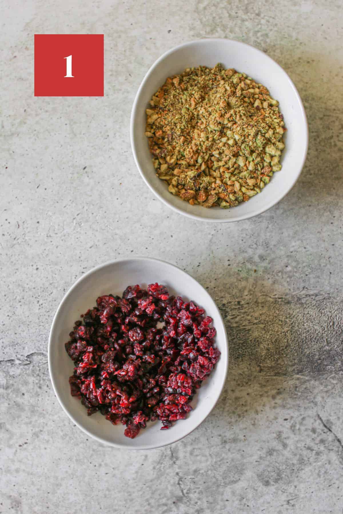 Two small white low bowls. One with crushed and finely ground pistachios and the other with diced dried cranberries. The bowls sit on a cement background. In the upper left corner is a dark red square with a white '1' in the center.