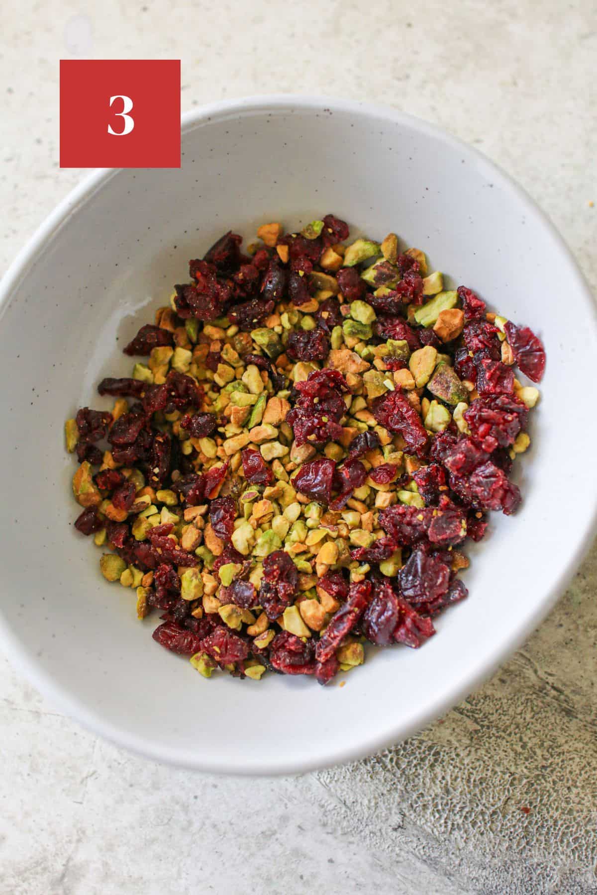 Dried cranberries and chopped pistachios combined in a white bowl on a cement background. In the upper left corner is a dark red square with a white '3' in the center.