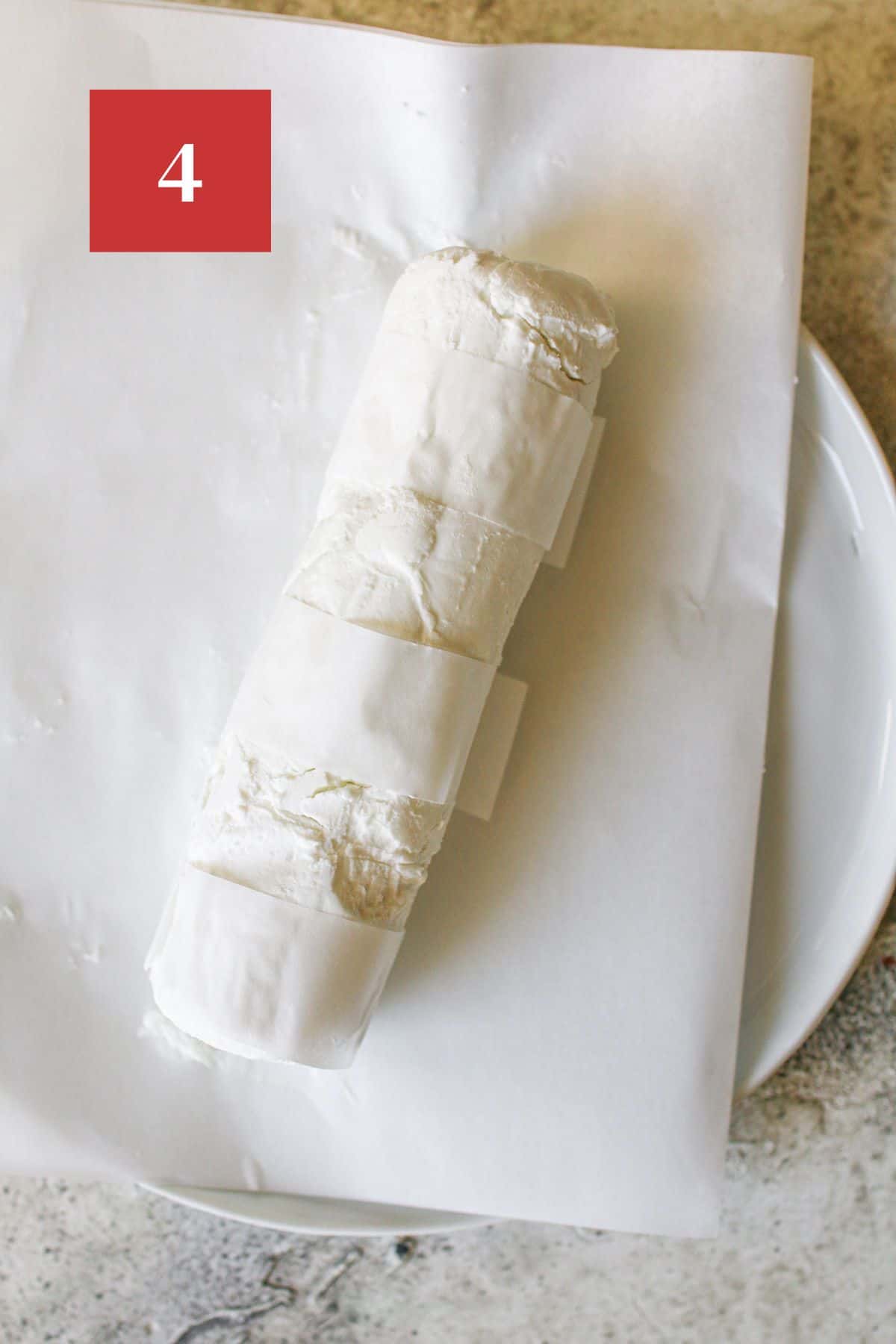 A goat cheese log on a white parchment paper on a white plate. The goat cheese it wrapped with strips of white parchment paper with some of the goat cheese exposed. The plate sits on a cement background. In the upper left corner is a dark red square with a white '4' in the center.