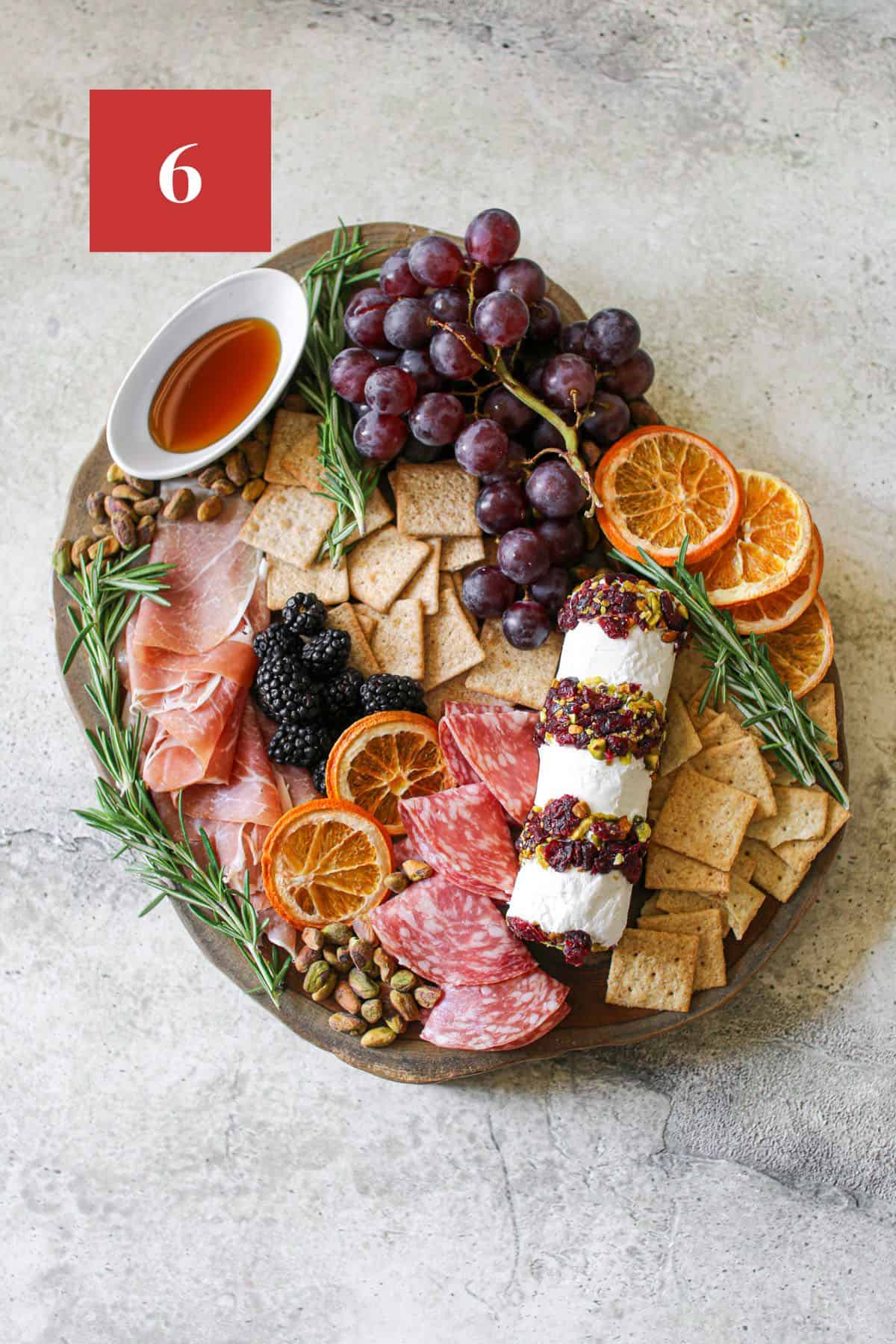 Overhead of a charcuterie board with on a wood trunk platter with a Cranberry Pistachio Cheese Log surrounded by crackers, grapes, meats and garnished with dried oranges and fresh rosemary. It sits on a stone background. In the upper left corner is a dark red square with a white '6' in the center.