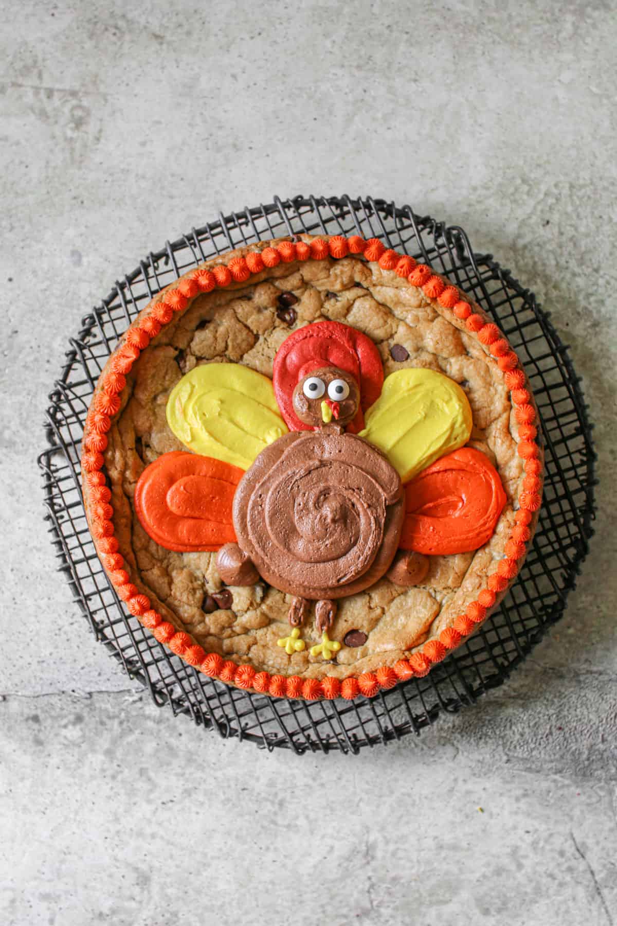 Turkey Cookie Cake with a buttercream turkey with an orange buttercream border on black wire trivet on a stone background.
