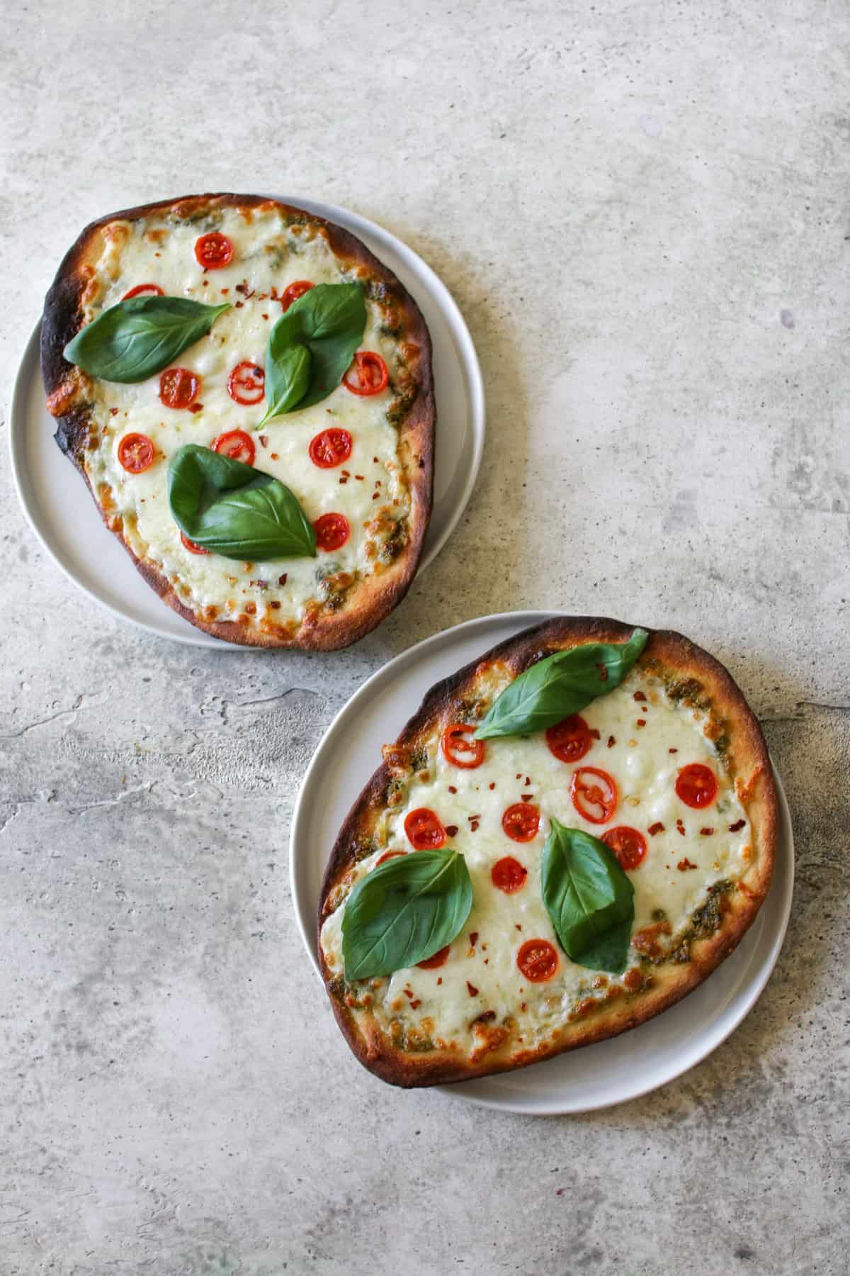 Two Pesto Naan Plates on white lates on a cement background. On each naan pizza is topped 3 basil leaves, thinly sliced tomatoes and chili flakes.