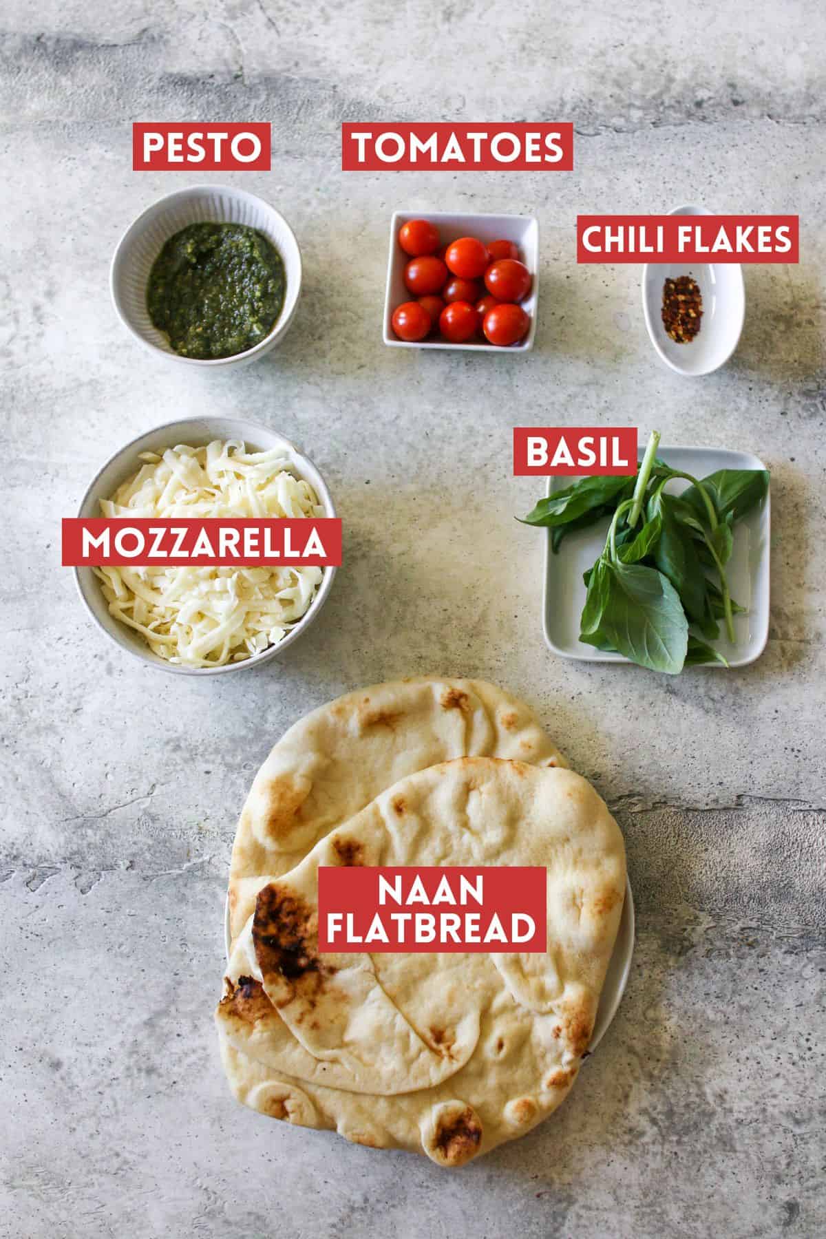 Pesto Naan pizza ingredients separated into individual white bowls or plates. Each item is labeled with a dark red rectangle box with white text in all caps. Everything sits on a cement background.
