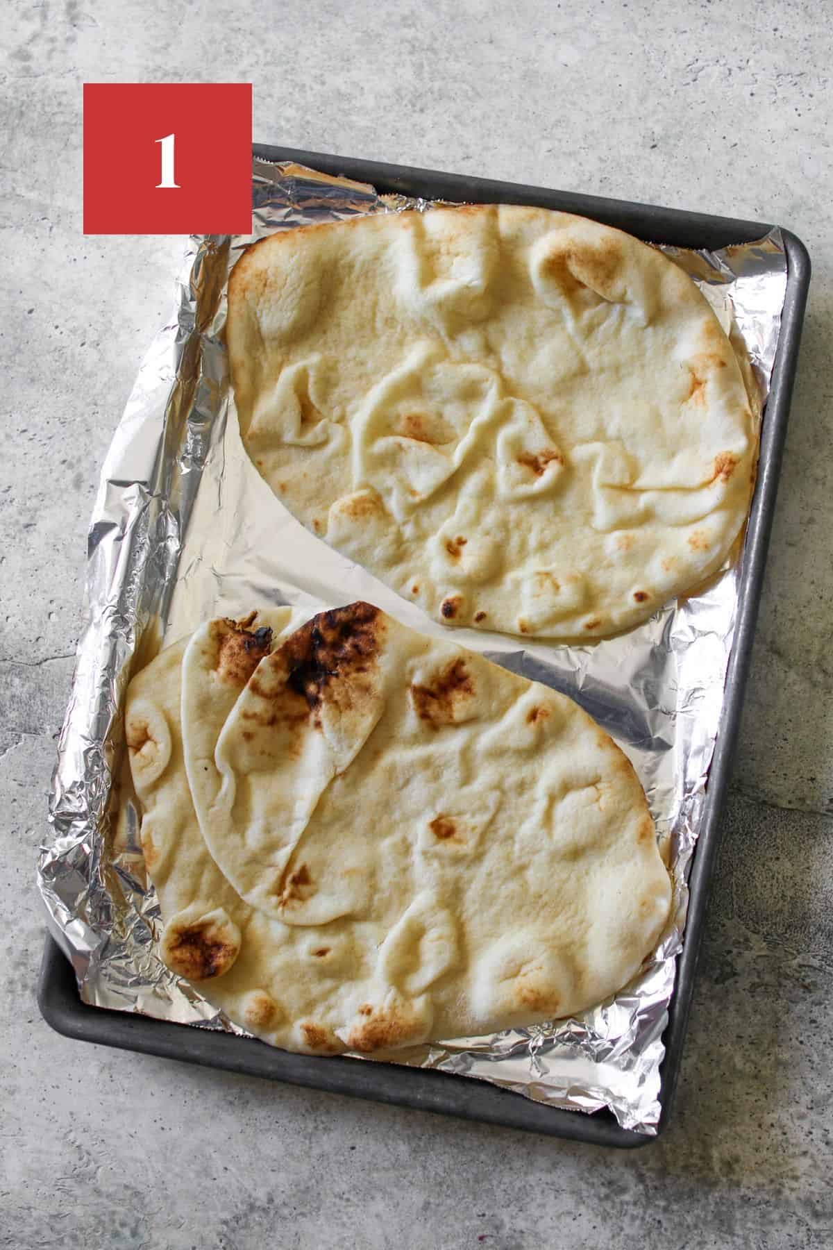 2 pieces of naan on a baking sheet lined with aluminum foil on a cement background. In the upper left corner is a dark red square with a white '1' in the center.