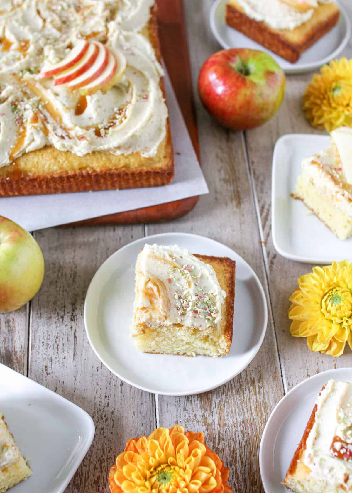 Sliced vanilla cake on small white plates. Each is topped with apple caramel frosting. The plates sit on a wood plank background with whole apples and yellow/orange flowers.