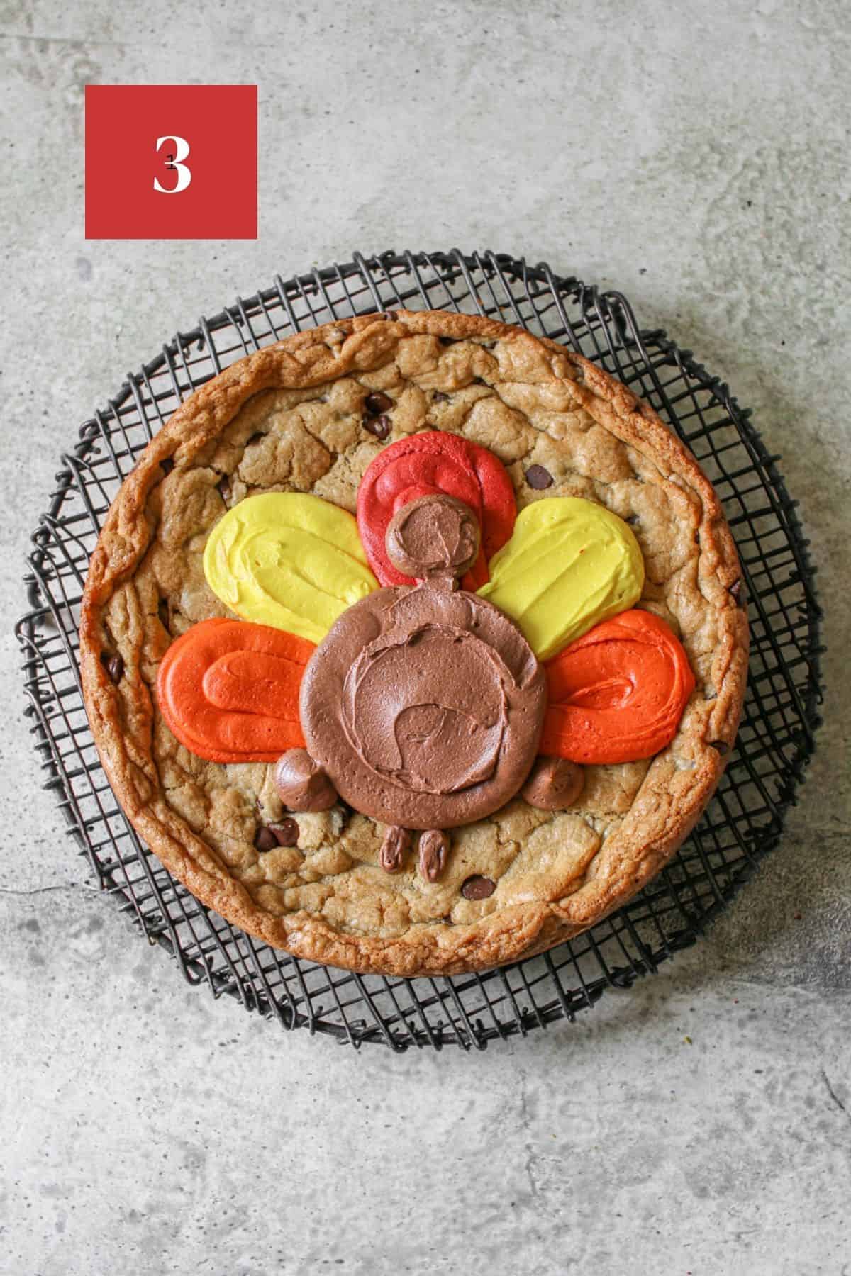 A cookie cake on a black wire trivet on a stone background. The cookie cake has 5 feathers that are tear drop shaped. From left to right, the feathers are orange, yellow, red, yellow and orange. On top of the feathers, is a brown turkey body, head and wings, neck and legs on the side. In the upper left corner is a dark red square with a white '3' in the center.