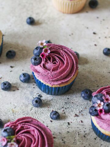 Blueberry Buttercream frosted vanilla cupcakes on a cement background surrounded by fresh blueberries.