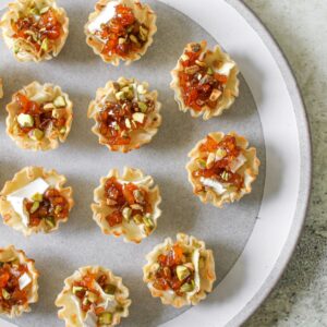 Apricot Brie Bites on a grey platter with a white ring. The platter sits on a stone background. Each Apricot Brie Bite is made of a phyllo cup as the base, melted brie, an apricot topping and crushed pistachios.
