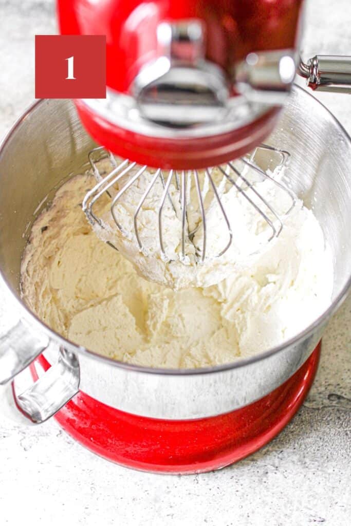 A stand mixer with whipped cream to stiff peaks. The red stand mixer has a whisk attachment and sits on a stone background. In the upper left corner is a dark red square with a white '1' in the center.