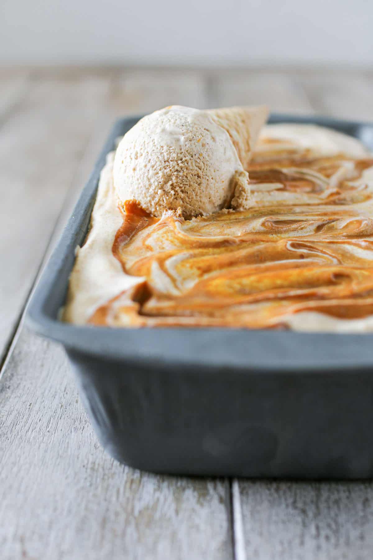 Side view of metal loaf pan with No Churn Pumpkin Ice Cream with a scoop on a sugar cone on top of the ice cream. The loaf pan sits on a wood plank background.