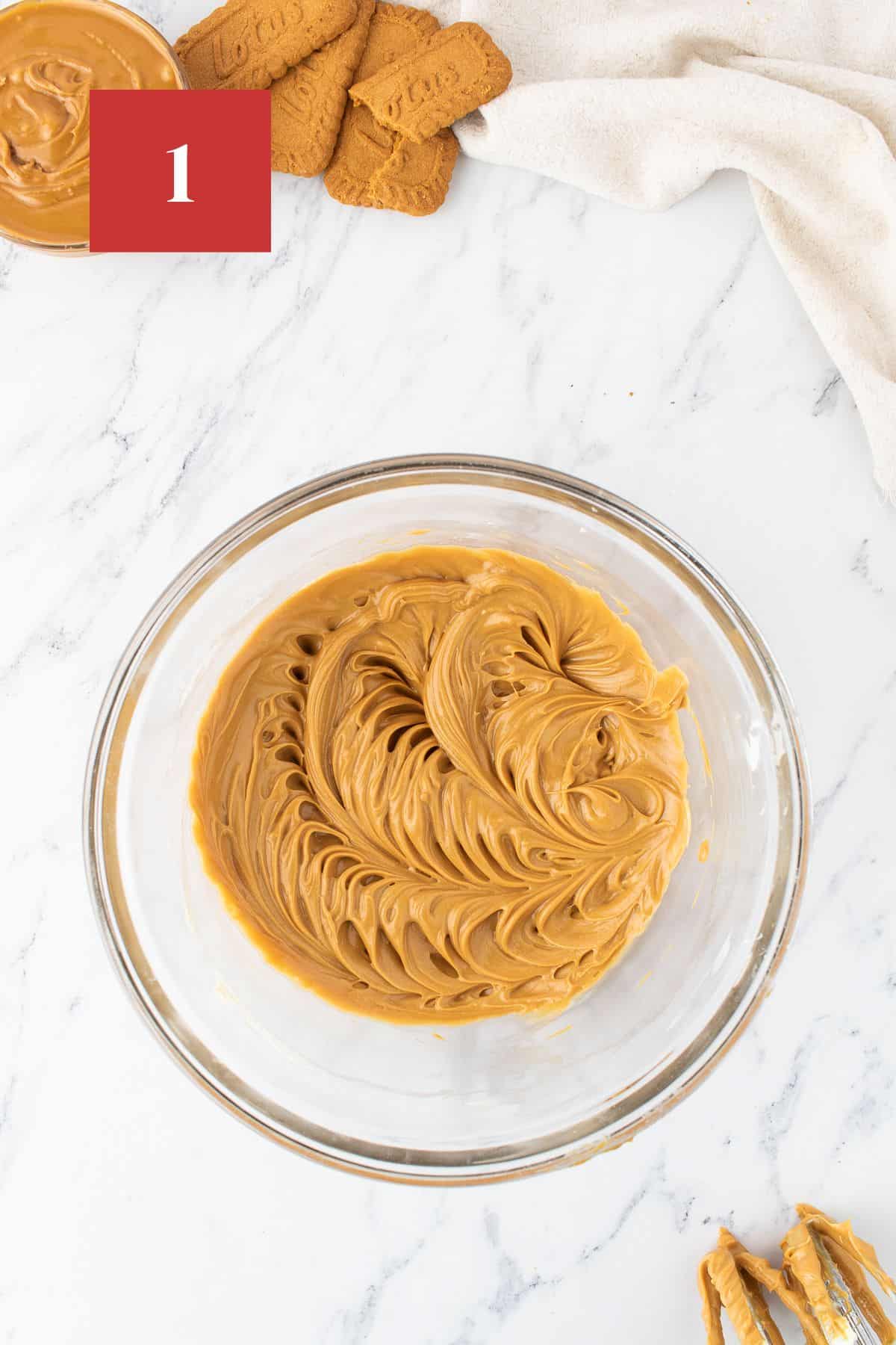 Whipped biscoff and butter in a glass bowl on a white marble background. . In the upper left corner is a dark red square with a white '1' in the center.