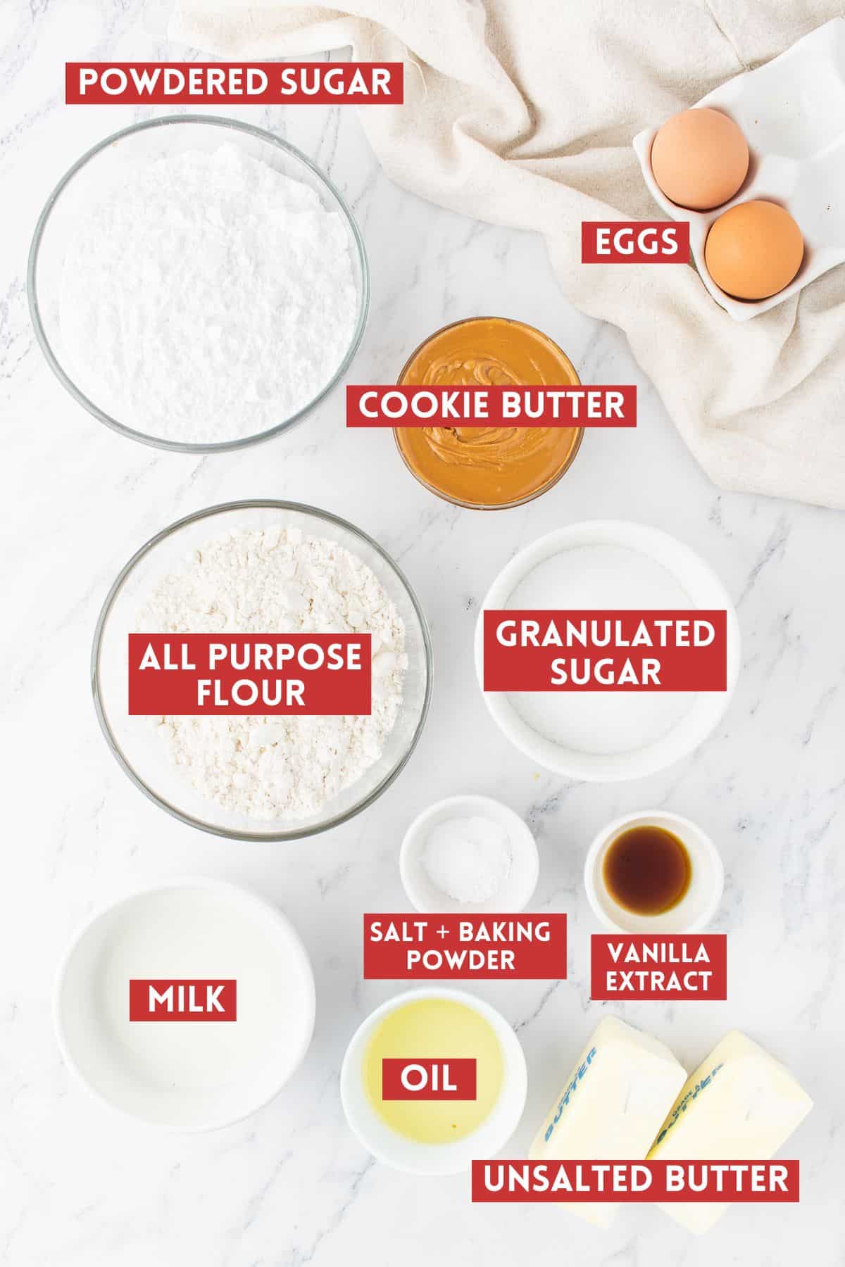Biscoff Cupcake ingredients on a white marble background. Each ingredient is labeled with a dark red rectangle with white text in all capitalization. 