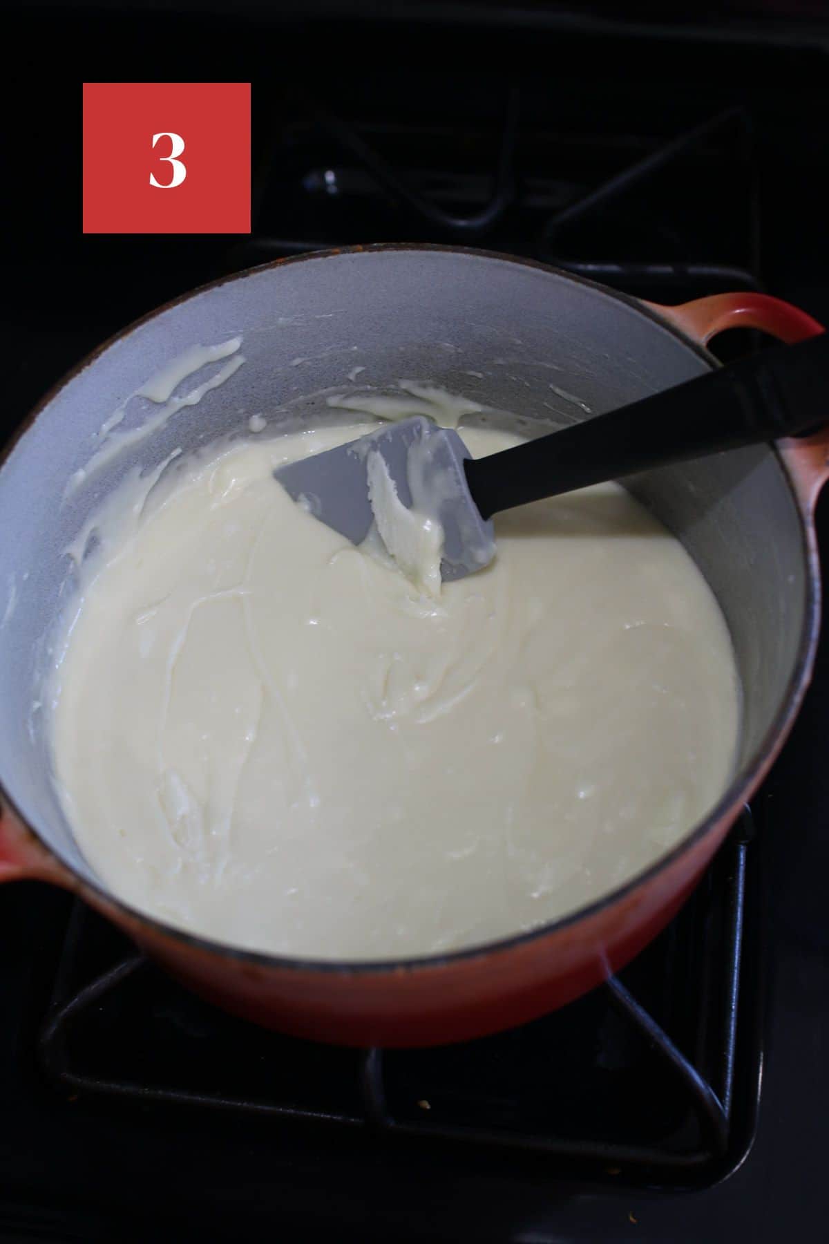 A small red dutch oven on top of the stove with white chocolate fudge mixtures and a silicone spatula. In the upper left corner is a dark red square with a white '3' in the center.