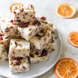 White chocolate Cranberry Pistachio Fudge stacked on top of each other on a white plate on a cement background and a couple of dried orange slices are spread around.