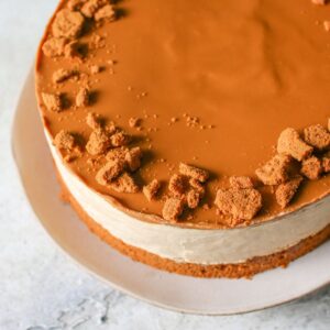 Whole No Bake Biscoff Cheesecake on a light brown plate on a cement background. The cheesecake has a biscoff crust and cookie butter topping with crushed cookies on top.