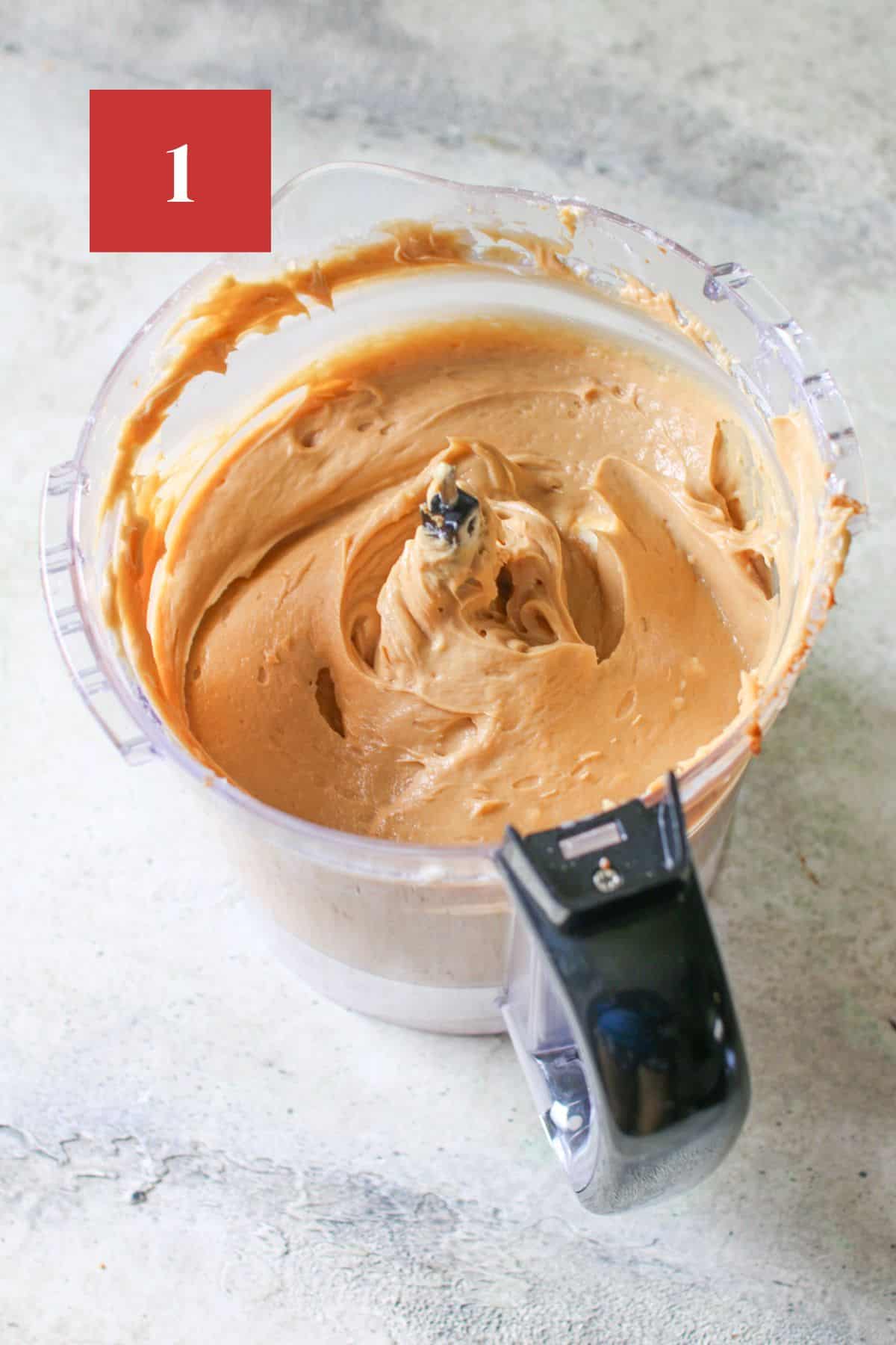 A large food processor with top removed, in the container is a cheesecake biscoff filling. it sits on a cement background. In the upper left corner is a dark red square with a white '1'. 