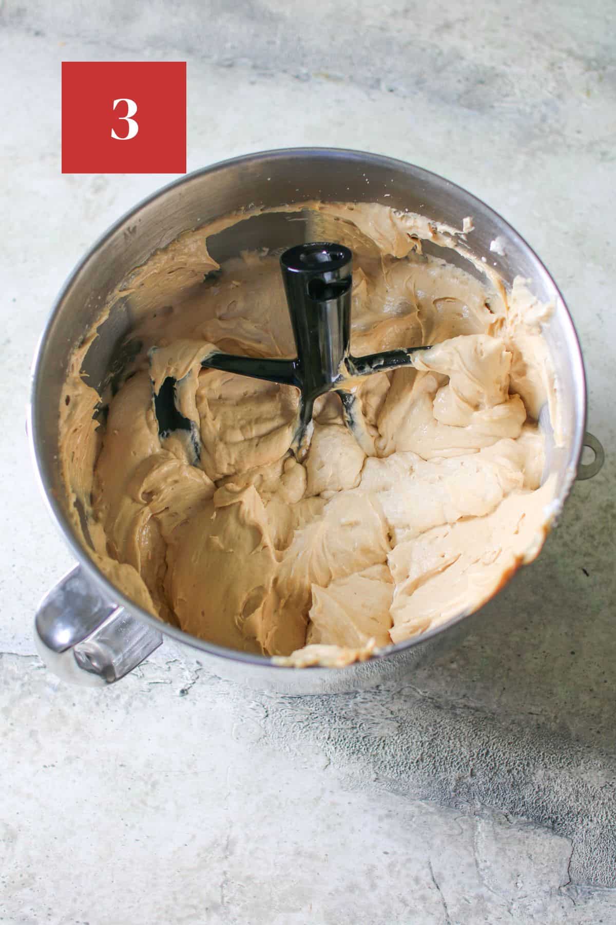 Biscoff cheesecake filling in a metal mixing bowl with a  a beater attachment on top. The bowl sits on a cement background.  In the upper left corner is a dark red square with a white '3'. 