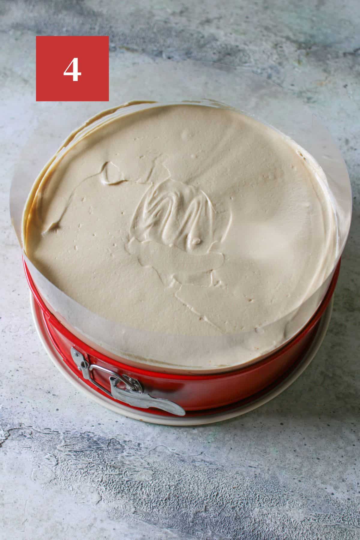 No bake biscoff cheesecake in a red springform pan with acetate sheet on a concrete background. In the upper left corner is a dark red square with a white '4'. 