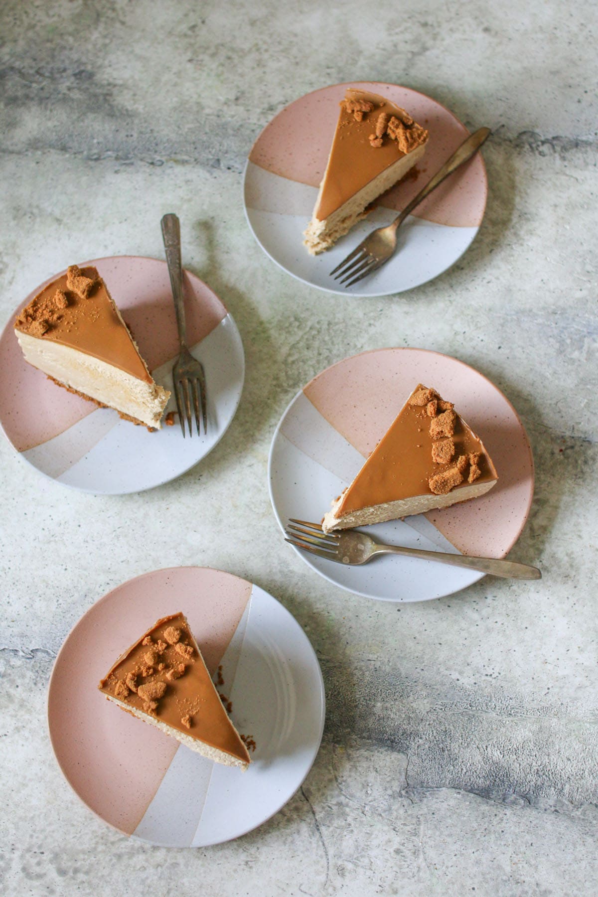 4 slices of No Bake Biscoff Cheesecake on pink and white plates on a cement background. 3 plates had an antique fork.