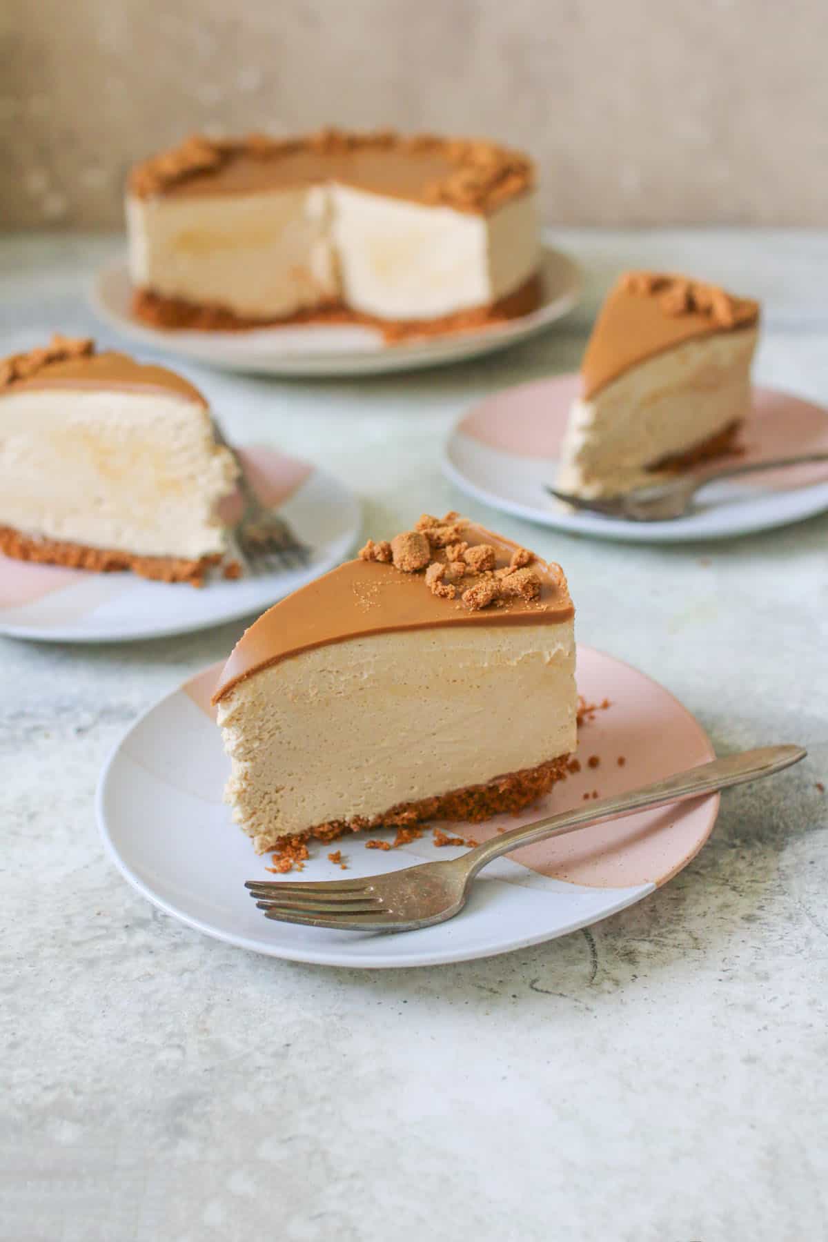 Close up of sliced No Bake Biscoff Cheesecake with more slices in the back with the rest of the cheesecake. A slice of cheesecake on a pink white plate with an antique fork on top.