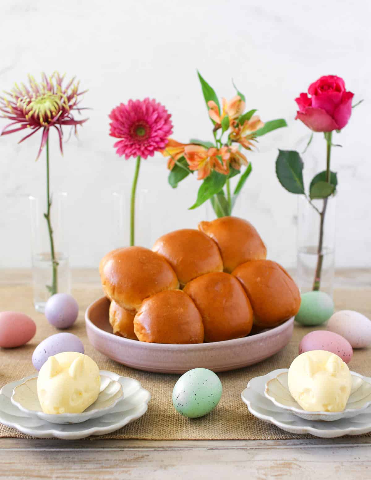 Two Butter shaped like a bunny sits on a small scalloped plate on top of a slightly larger scallop plate. Behind it is a bowl of bread rolls. Behind the bread are four thin clear glass vases each with a flower and its surrounded by pastel eggs.