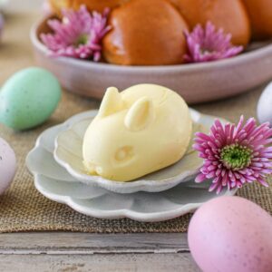 Butter shaped like a bunny sits on a small scalloped plate on top of a slightly larger scallop plate. Behind it is a bowl of bread rolls. Its surrounded by pink flowers and pastel eggs.