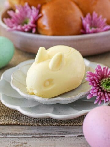 Butter shaped like a bunny sits on a small scalloped plate on top of a slightly larger scallop plate. Behind it is a bowl of bread rolls. Its surrounded by pink flowers and pastel eggs.