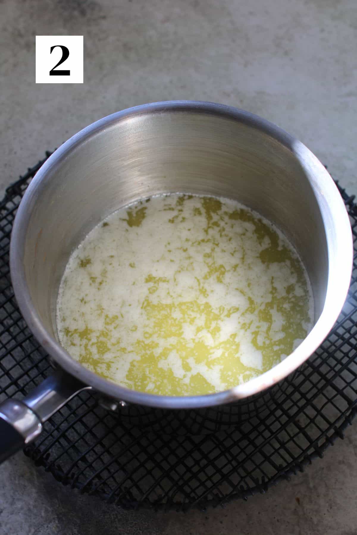 A saucepan with melted butter and lemon zest sits on a black wire trivet on a cement background. In the upper left corner, is a white square with a black number '2'.