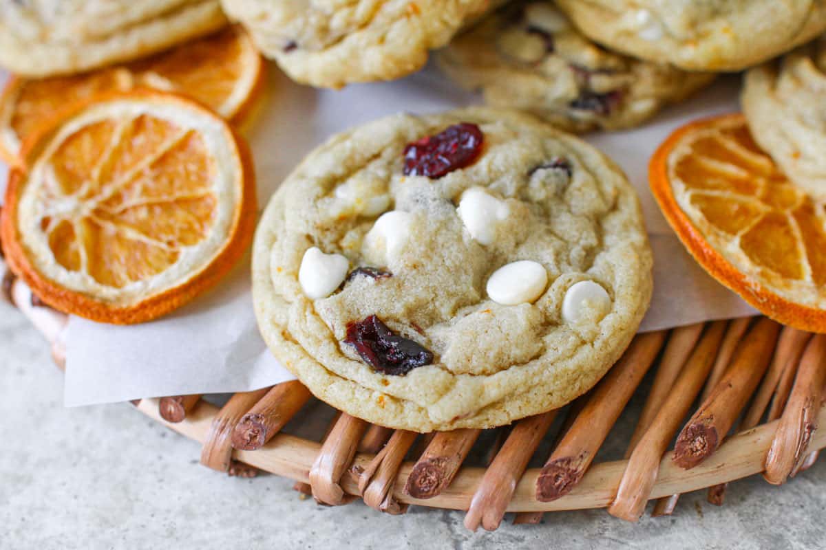 Close up on a High Altitude White Chocolate Cranberry Orange Cookie on a rattan tray with dried orange slices and more cookies in the background.