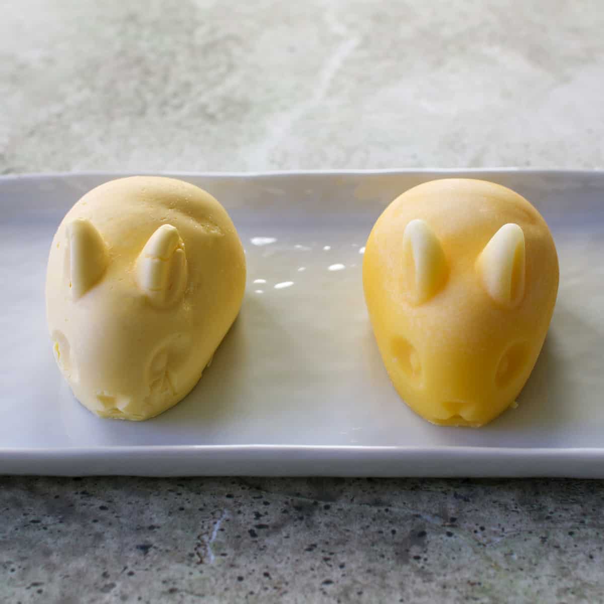 Two butter bunnies on a white rectangle plate. The bunny on the left is made of softened butter and the right bunny is made of melted butter.