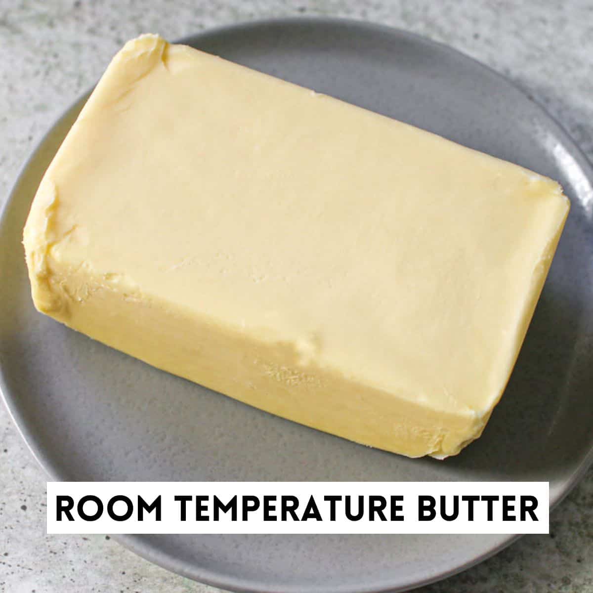 A large rectangle piece of butter on a small grey plate sitting on a cement background. On the bottom is a white rectangle with the words 'ROOM TEMPERATURE BUTTER" in black font.