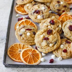 High Altitude White Chocolate Cranberry Orange Cookies on a vintage baking sheet lined with white parchment paper. The cookies are scattered along with dried sliced oranges and white chocolate chips and dried cranberries.