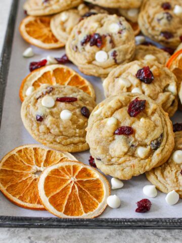 High Altitude White Chocolate Cranberry Orange Cookies on a vintage baking sheet lined with white parchment paper. The cookies are scattered along with dried sliced oranges and white chocolate chips and dried cranberries.