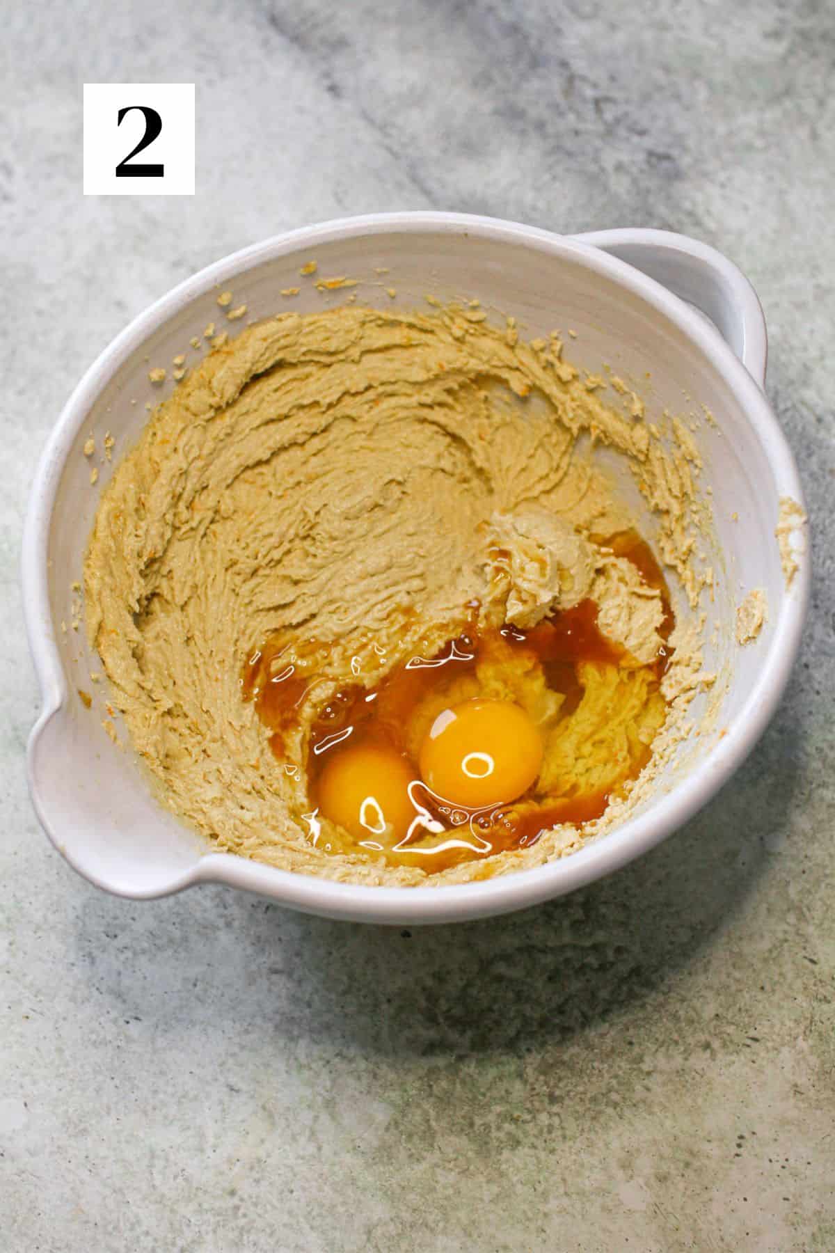 A wet cookie dough in a white ceramic mixing bowl with vanilla extract and two eggs. The bowl sits on a cement background. In the upper left corner is a white square with a black '2'.