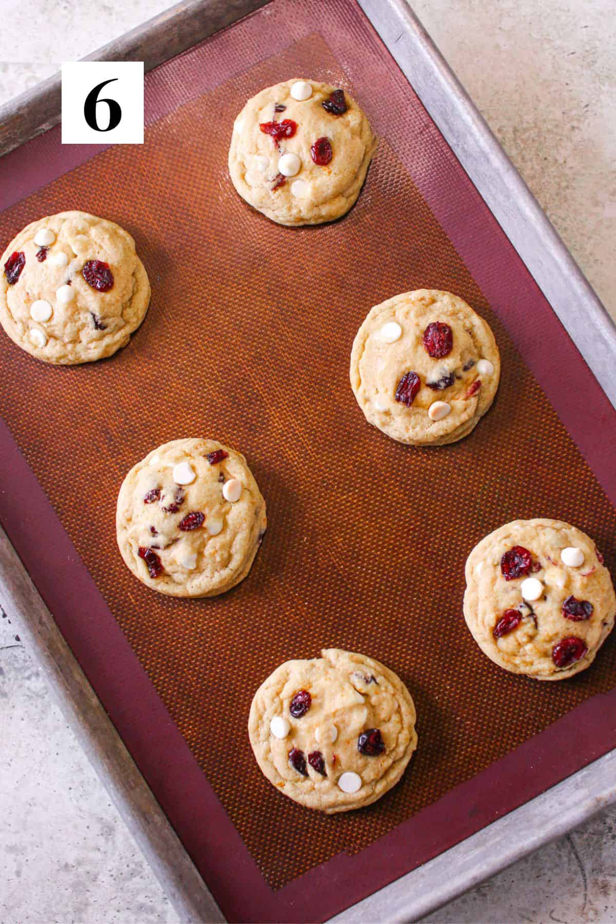 Six High Altitude White Chocolate Cranberry Orange Cookies on a silicone mat on a baking tray. Each cookie dough ball is topped with extra dried cranberries and white chocolate chips. In the upper left corner is a white square with a black '6'.