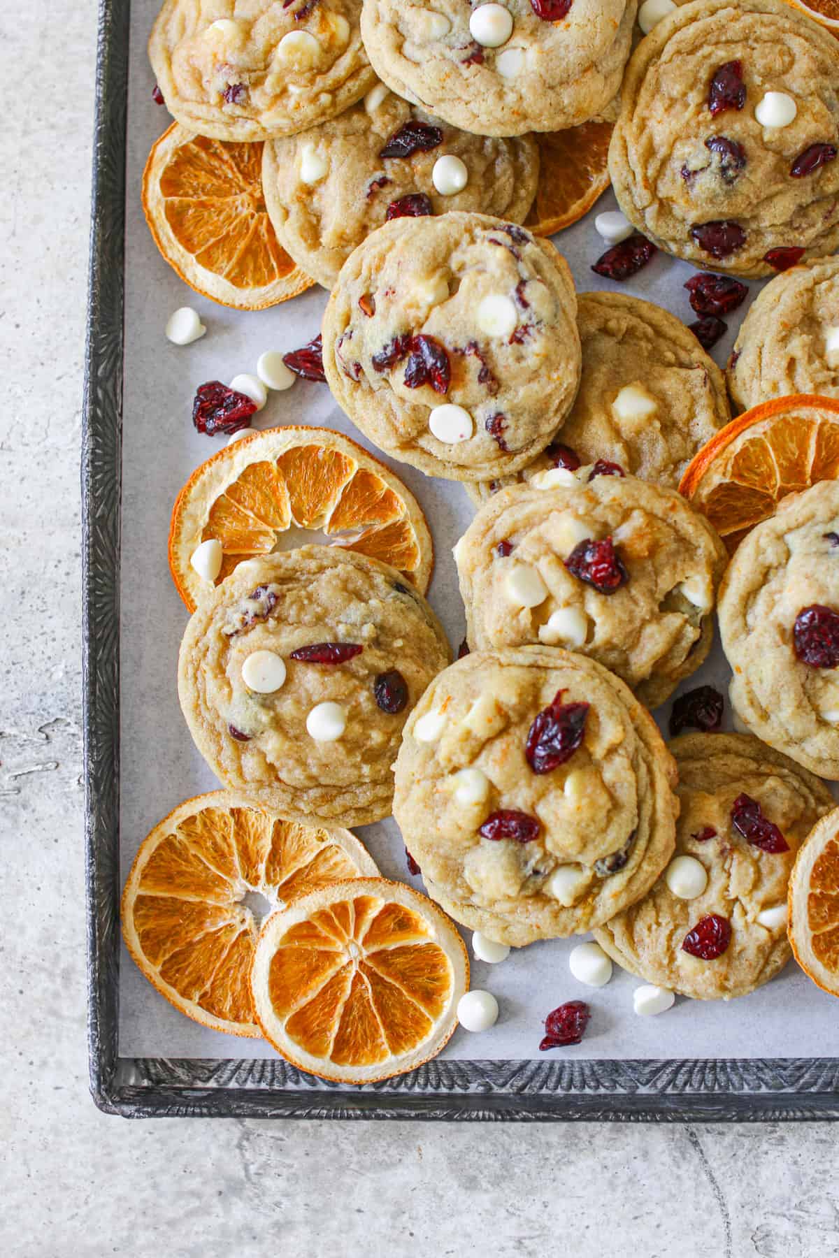 Overhead of High Altitude White Chocolate Cranberry Orange Cookies on a vintage baking sheet lined with white parchment paper. The cookies are scattered along with dried sliced oranges and white chocolate chips and dried cranberries.